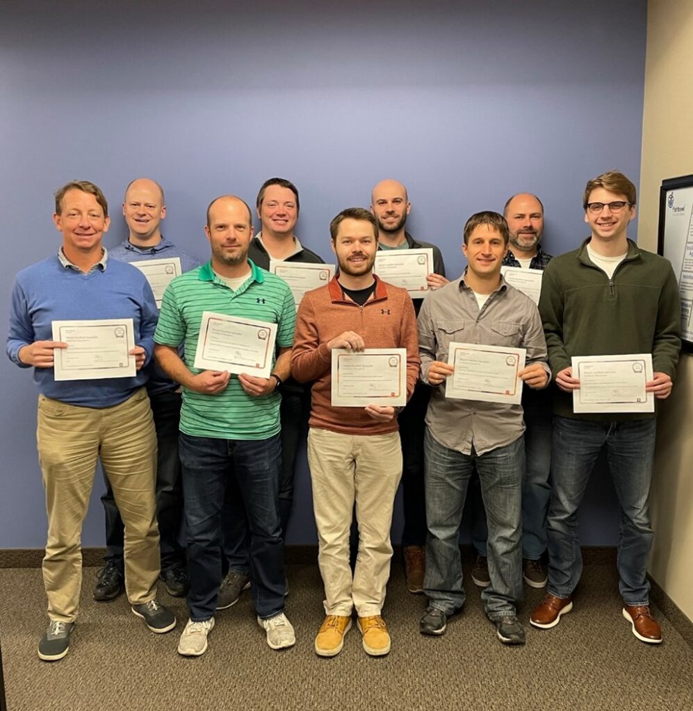 Nine Fishbowl Solutions employees stand against a blue wall holding their Oracle Cloud certifications